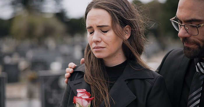Grieving woman holding a rose, comforted by a man at a cemetery near a vandalized daughter's grave.