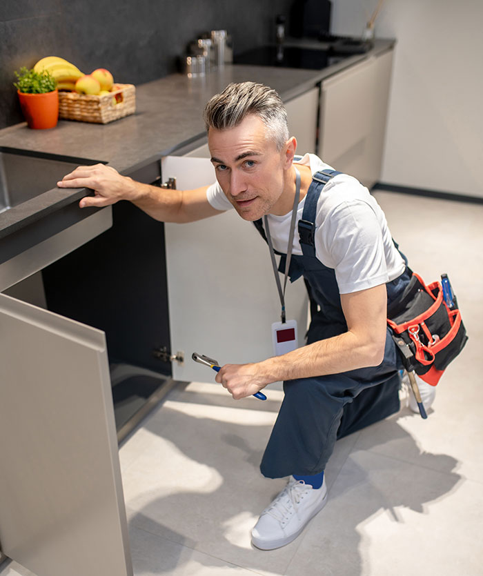 Man in work overalls kneeling by kitchen cabinet, holding a wrench with a suspicious expression, depicting a creep scenario.