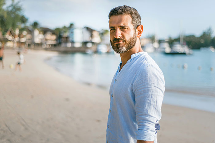 Man standing on a sunny beach, wearing a light blue shirt, representing men hitting on women like creeps.