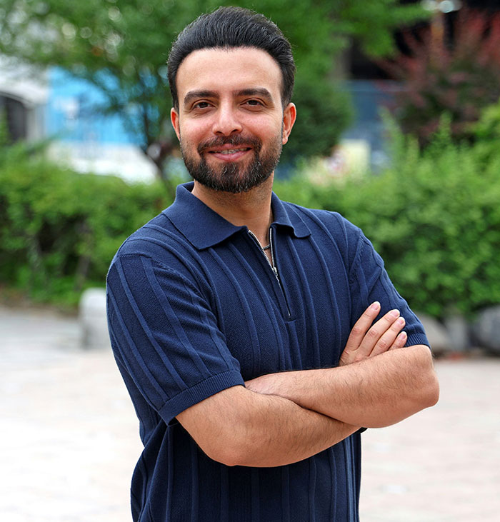 Man with beard and mustache wearing a navy blue shirt standing outdoors with arms crossed and smiling confidently.