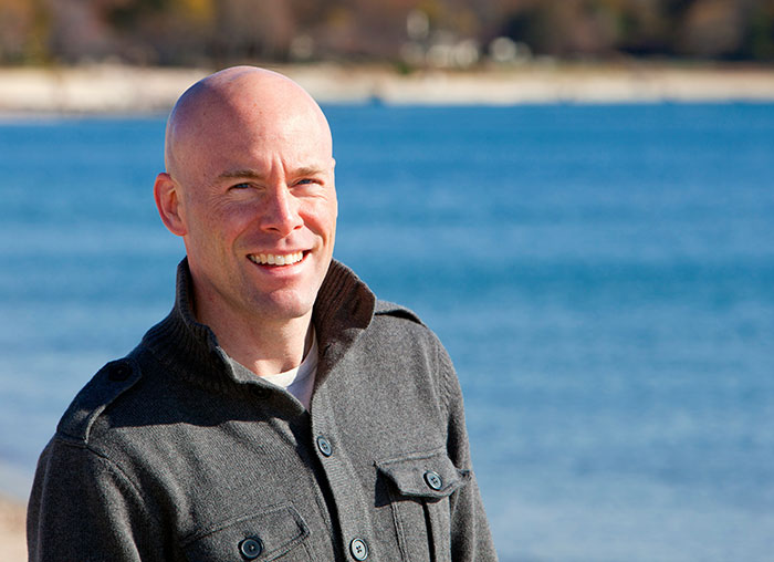 Smiling bald man wearing a buttoned gray jacket standing outdoors near water in a casual setting.