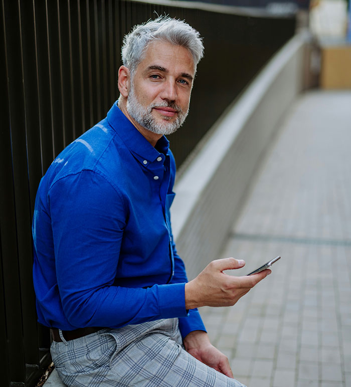 Middle-aged man in a blue shirt sitting outdoors holding a phone, representing men hitting on women like creeps stories.