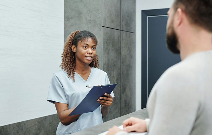 Young woman in scrubs holding clipboard talking to a man, illustrating stories of men hitting on women like creeps.