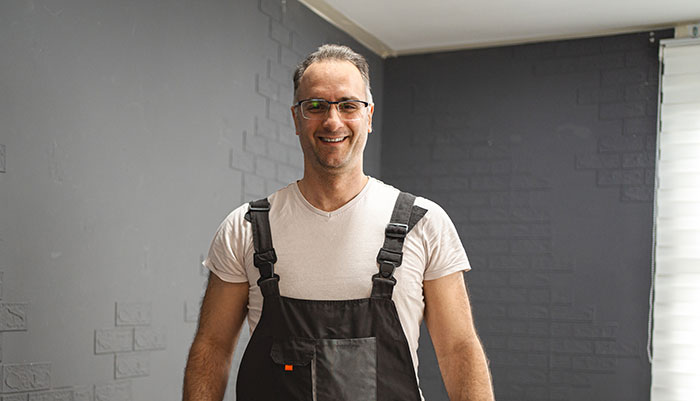 Man wearing glasses and overalls smiling indoors, relating to stories of men hitting on women like creeps.