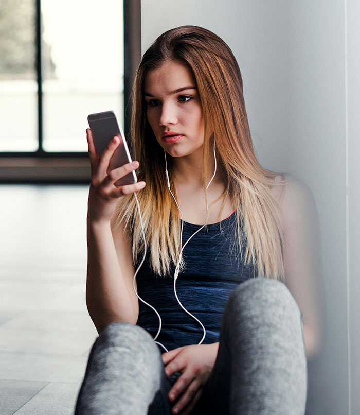 Young woman sitting against a wall, wearing earphones and looking at her phone, reflecting stories of men hitting on women.