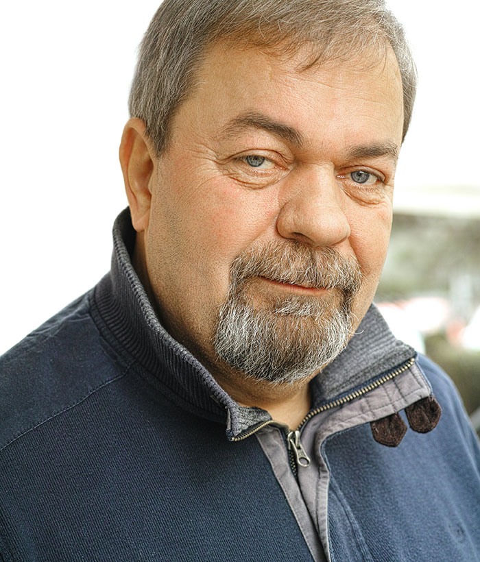 Close-up portrait of a middle-aged man with gray beard and blue eyes wearing a navy zip-up sweater outdoors.