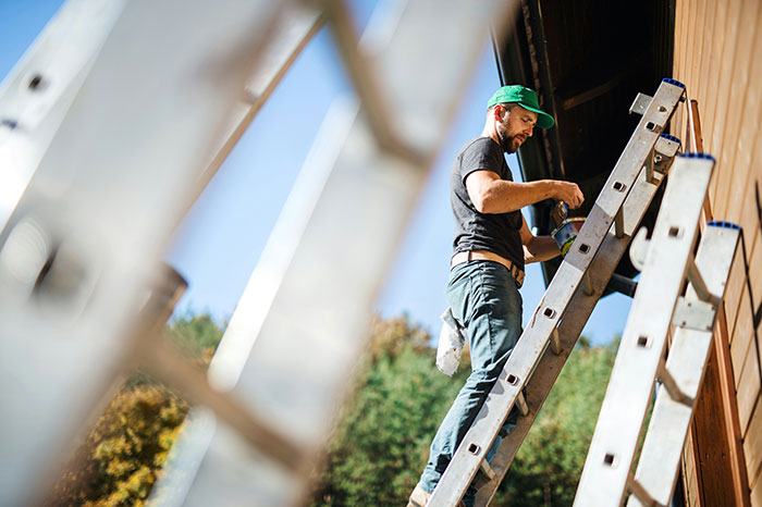 Man wearing green cap and black shirt on ladder, painting a wooden wall outdoors, illustrating stories of men hitting on women.