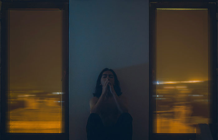 Woman sitting alone at night between two windows with yellow city lights, reflecting feelings of discomfort and creepiness.