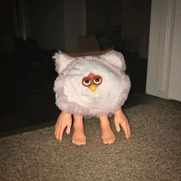 Weird and cringey photo of a Furby toy with human hands and feet wearing a cowboy hat on a carpeted floor.