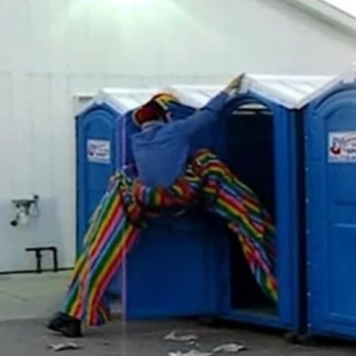 Person wearing rainbow-striped pants awkwardly squeezed halfway inside a portable toilet, creating a weird and cringey photo moment.