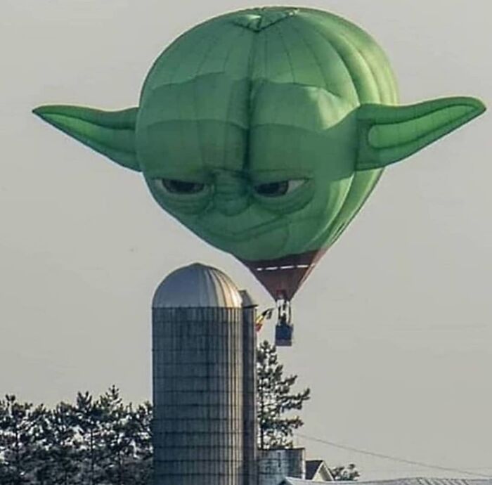 Hot air balloon shaped like a large green head with ears flying near a silo in a weird and cringey photo.