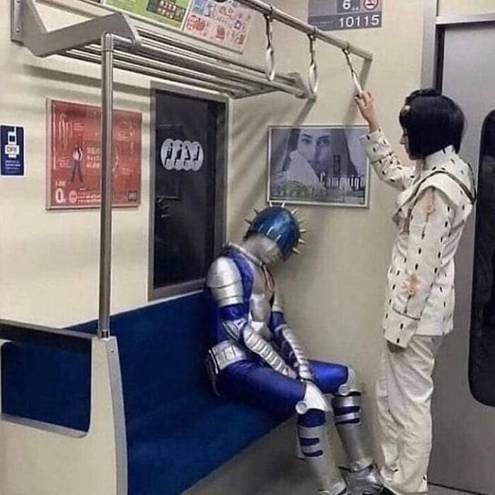 Person in futuristic armor sitting on subway bench looking down while another person stands holding a strap in a weird and cringey photo