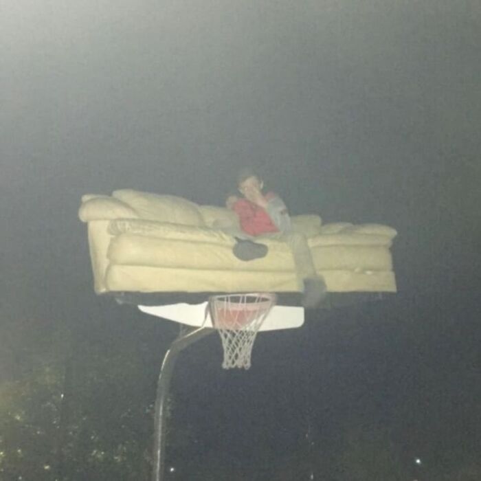 Person sitting on a couch oddly placed on top of a basketball hoop, a weird and cringey photo difficult to look at.