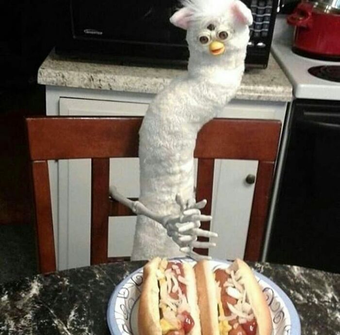 Weird and cringey photo of a distorted Furby doll with skeletal hands sitting at a table with hot dogs.