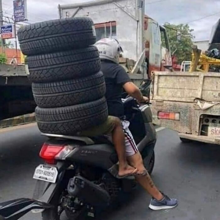 Man riding a scooter carrying an unsafe stack of tires, a weird and cringey photo difficult to look at on a busy street.