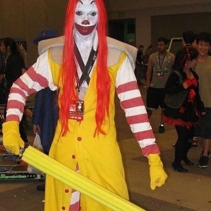 Person in a weird and cringey clown costume with red hair and yellow outfit holding a large foam bat at a convention.