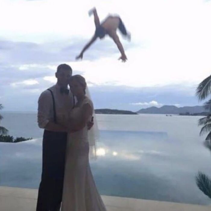A couple in wedding attire posing by a pool with a blurry figure awkwardly flipping in the background creating a weird and cringey photo.