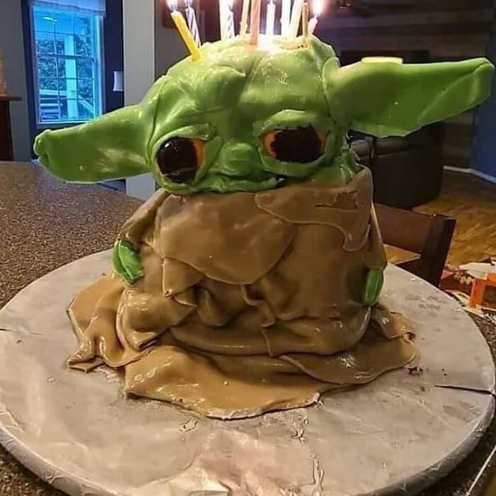 Cringey and weird homemade cake shaped like a green character with large ears and wrinkled brown clothing on a marble surface.