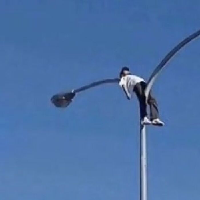 Person awkwardly hanging on a streetlight pole under a clear blue sky, a weird and cringey photo difficult to look at.