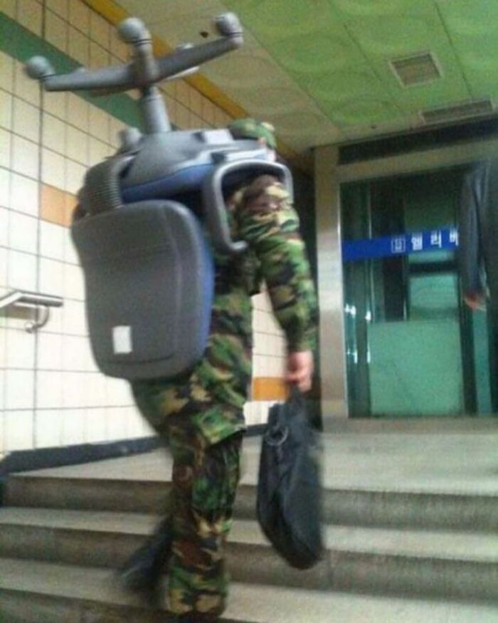 Person in camouflage carrying a large, unusual backpack shaped like an office chair in a hallway, weird and cringey photo.