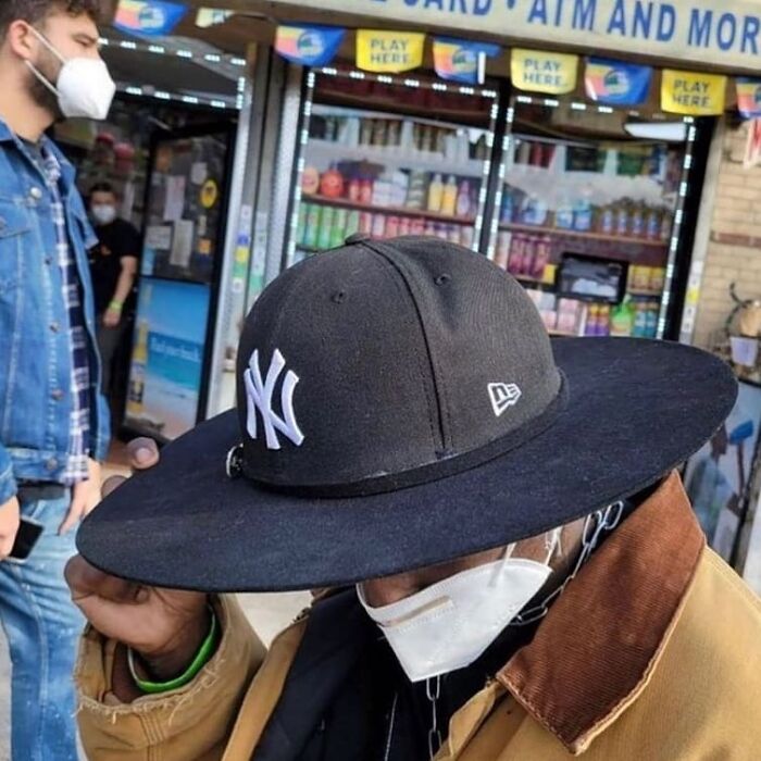 Person wearing an oversized New York Yankees cap and face mask outside a convenience store, a weird and cringey photo.