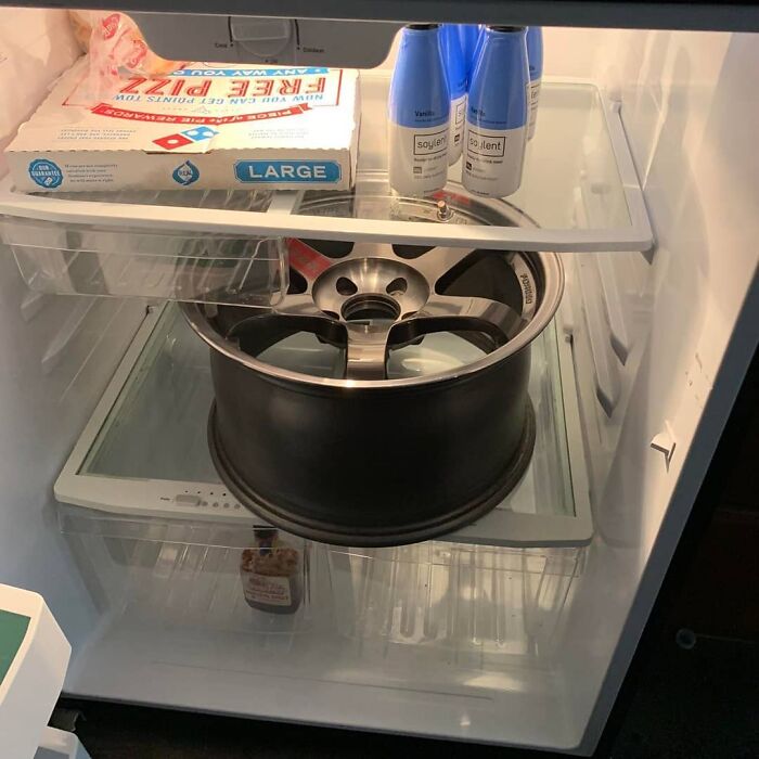 A car wheel rim stored inside a refrigerator alongside pizza and soylent drinks, showcasing a weird and cringey photo.