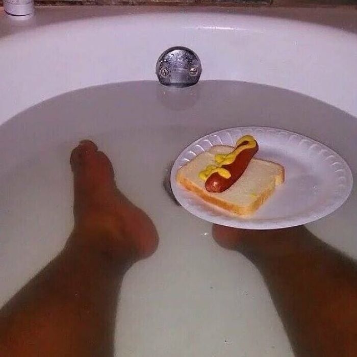 Foot soaking in a bathtub with a paper plate floating, holding a hot dog on bread with mustard, a weird and cringey photo.
