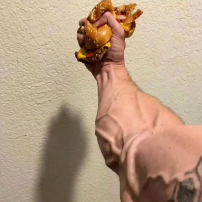 Muscular arm squeezing a messy cheeseburger, a weird and cringey photo difficult to look at.