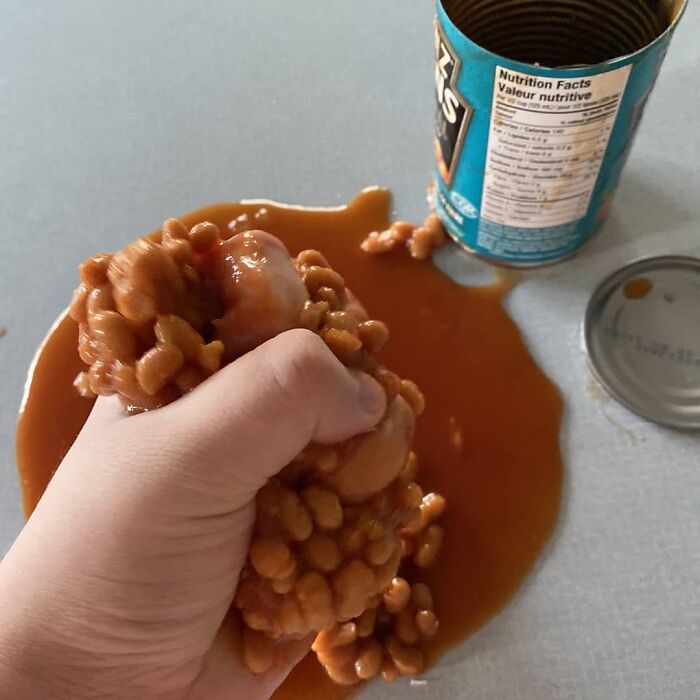 Hand squeezing spilled baked beans from an open can, creating a messy and cringey food scene difficult to look at.