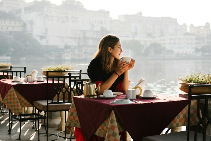 Woman enjoying tea at an outdoor café overlooking water, reflecting on truths sadly vanished from home countries.