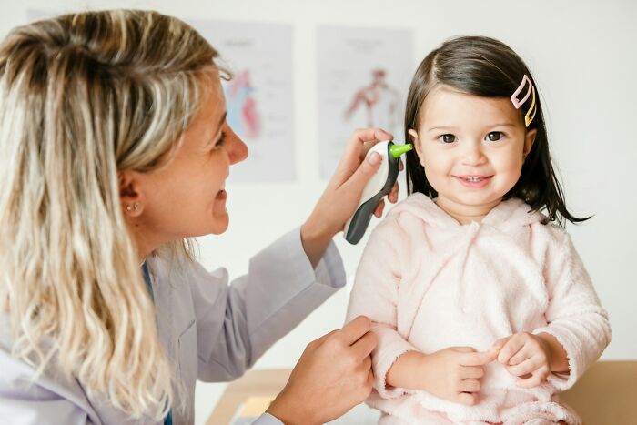 Doctor using an ear thermometer to check a smiling toddler in a medical exam room related to dumb ER visits brain malfunction.