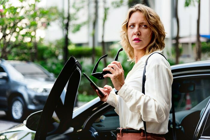 Woman holding sunglasses and phone by a car, showing strength and confidence against men’s creepy advances.