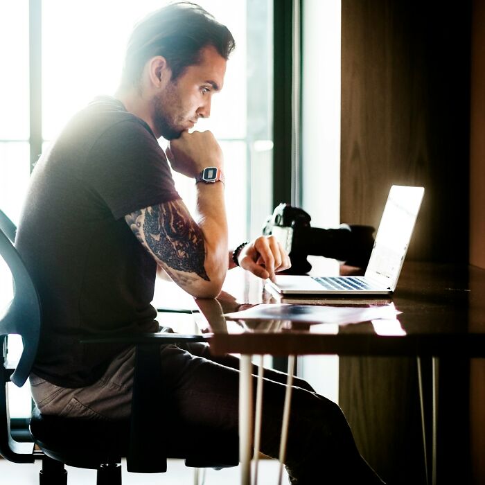 Man with tattooed arm researching interesting rabbit holes on laptop in bright, modern workspace with camera on desk.