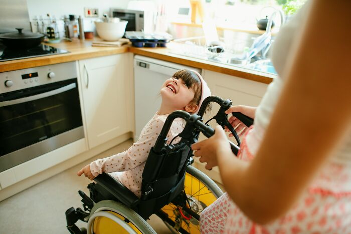 Young girl in wheelchair smiling in kitchen while caregiver pushes her, highlighting tough reasons in a relationship despite challenges.