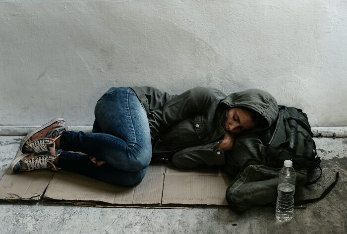Person in a hoodie curled up on cardboard, lying next to backpack and water bottle, illustrating times people destroyed their lives