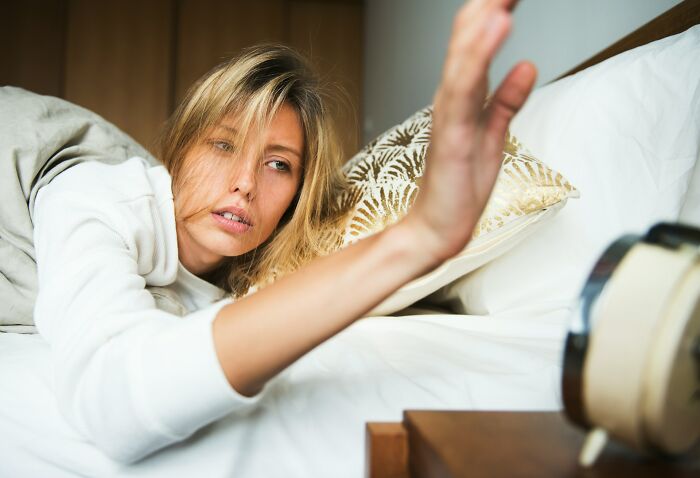 Tired woman reaching to turn off alarm clock in bed, illustrating a premium-tier zombie morning struggle concept.