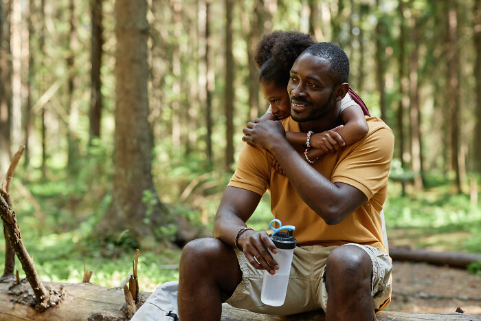 Man sitting on a log in the forest holding a water bottle while a child hugs him during a camping and hiking trip.