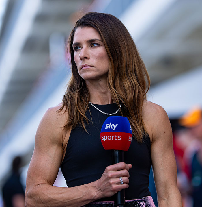 Danica Patrick holding a Sky Sports microphone, looking focused in a sleeveless black top at a racing event.