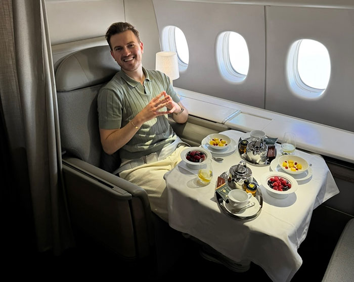 Man smiling while seated in first-class airplane cabin with gourmet meal, linked to influencer refusing to fly economy and stranded claim.