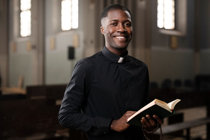 Young man in clergy attire holding a book and rosary beads inside a church, symbolizing dark industry secrets.