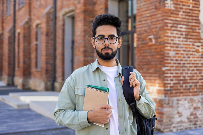 Young man with glasses holding notebook and backpack outside brick building, representing dark industry secrets shared by insiders.