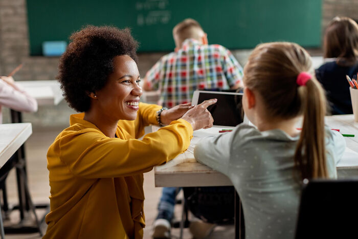 Teacher smiling and pointing at a tablet while explaining dark industry secrets to young students in a classroom setting.