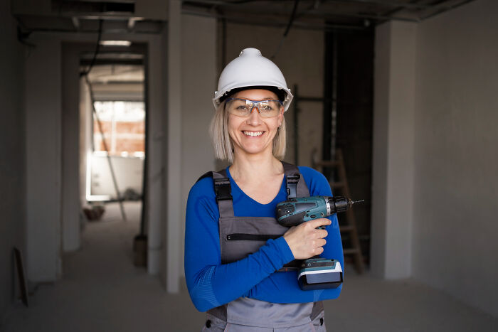 Woman construction worker wearing helmet and protective glasses holding a power drill inside unfinished building site, dark industry secrets concept.