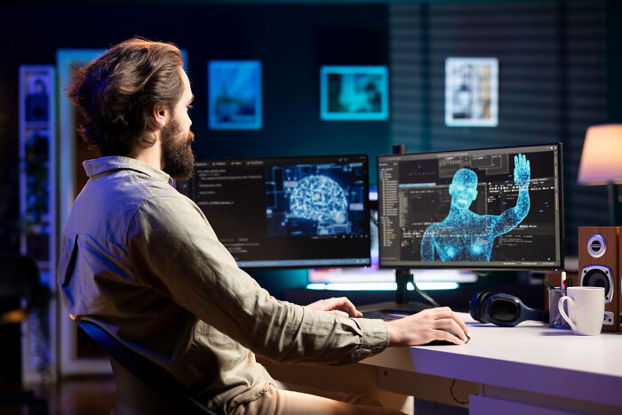 Man with beard working on dual monitors showing digital human and brain graphics, revealing dark industry secrets quietly.