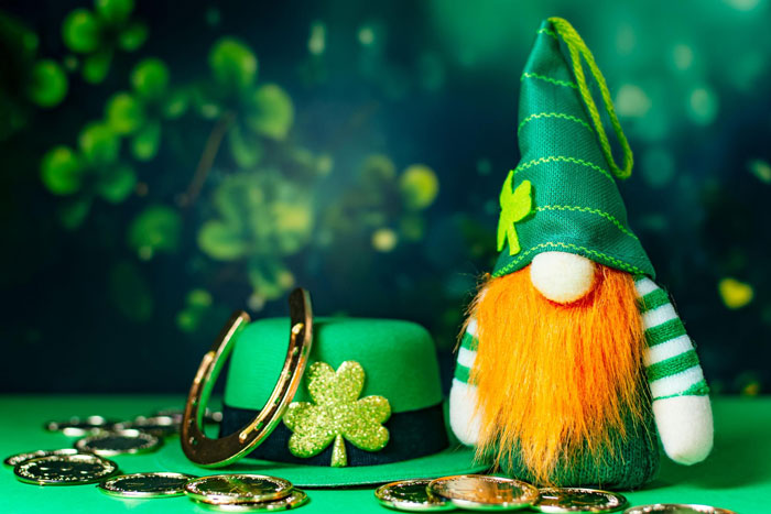 Green St. Patrick&rsquo;s Day hat, gold coins, horseshoe, and small gnome doll with orange beard on green surface.