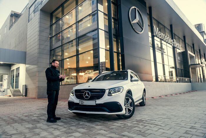 Man standing next to a white Mercedes-Benz outside a dealership, capturing a moment that feels like payback stories.
