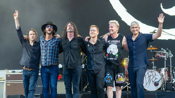 Six male musicians standing arm in arm on stage after a live concert in front of musical instruments and amplifiers