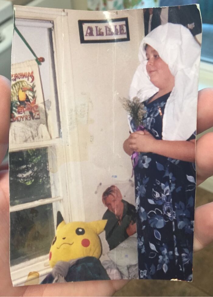 Child in a floral dress with a sheet on their head holding flowers in a hilariously awkward childhood pic with Pikachu plush.