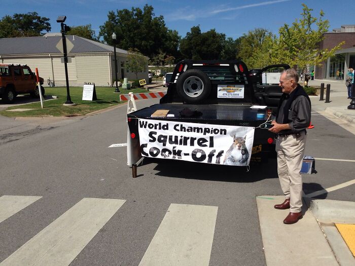 Man standing beside truck with World Champion squirrel cook-off banner at a street event capturing US state culture.