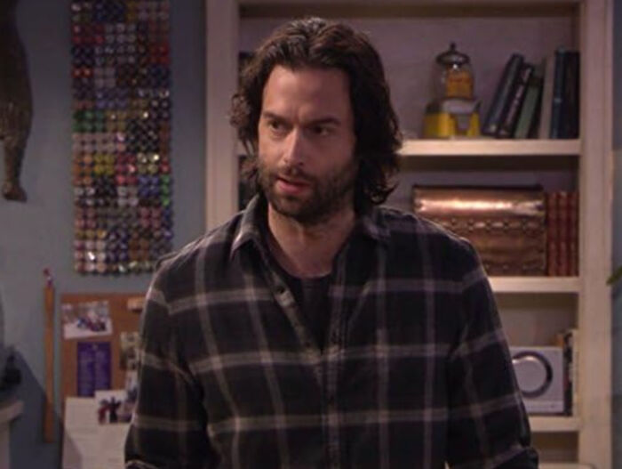 Man with long hair and beard wearing a plaid shirt, in a cozy room filled with shelves and various items, discussing celebrities.